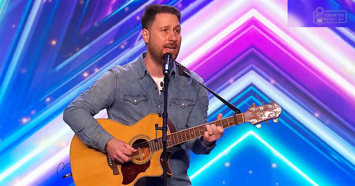 An adult male singer performing on a vibrant talent show stage with an acoustic guitar in hand, surrounded by dynamic lighting.