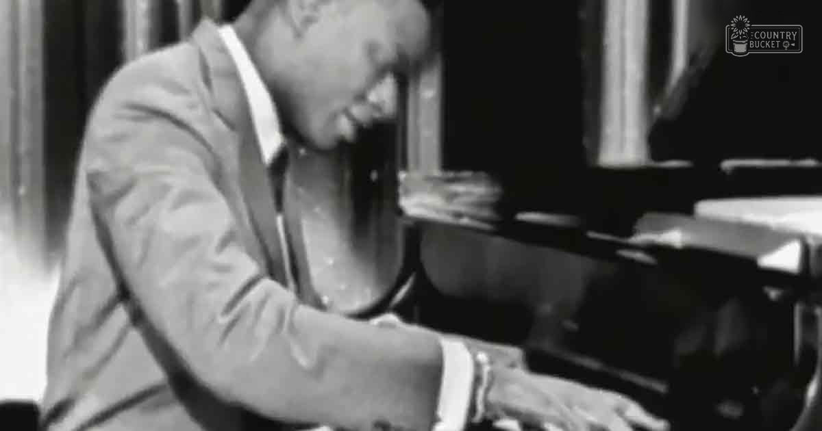 A young adult male in a light-colored suit jacket plays piano indoors with natural daylight streaming through curtains.