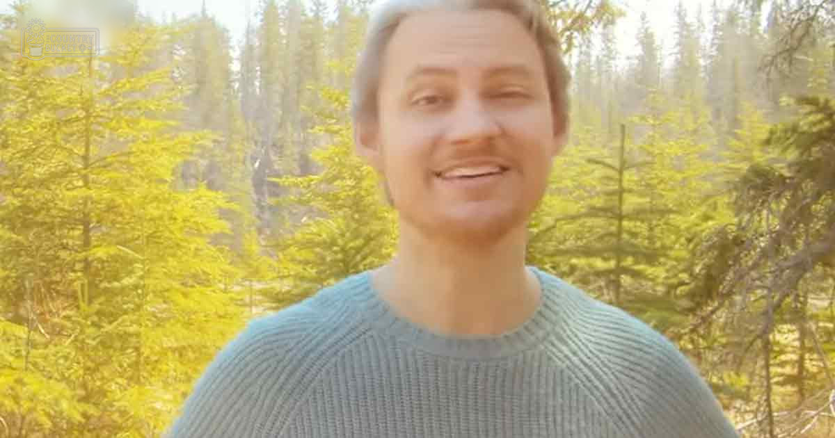 A young adult male wearing a light blue ribbed sweater in an outdoor forest setting with tall pine trees and soft golden light filtering through the foliage, suggesting early morning or late afternoon during spring or summer.