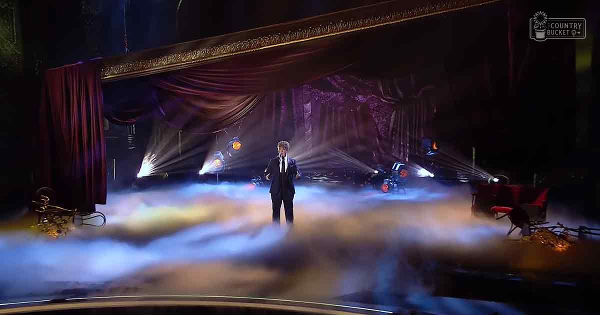 A young adult male performer in a dark suit and white shirt, surrounded by red curtains and stage lighting in an indoor entertainment venue.