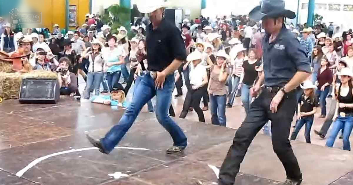 An outdoor dance performance with adults in cowboy attire, a speaker box with hay bales nearby, and a crowd gathered around the stage.