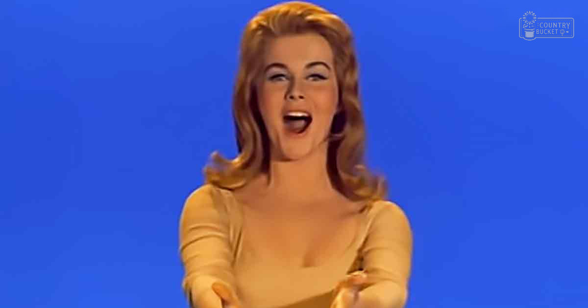 A young adult female wearing a mid-20th-century style, long-sleeved white top indoors against a solid blue backdrop.