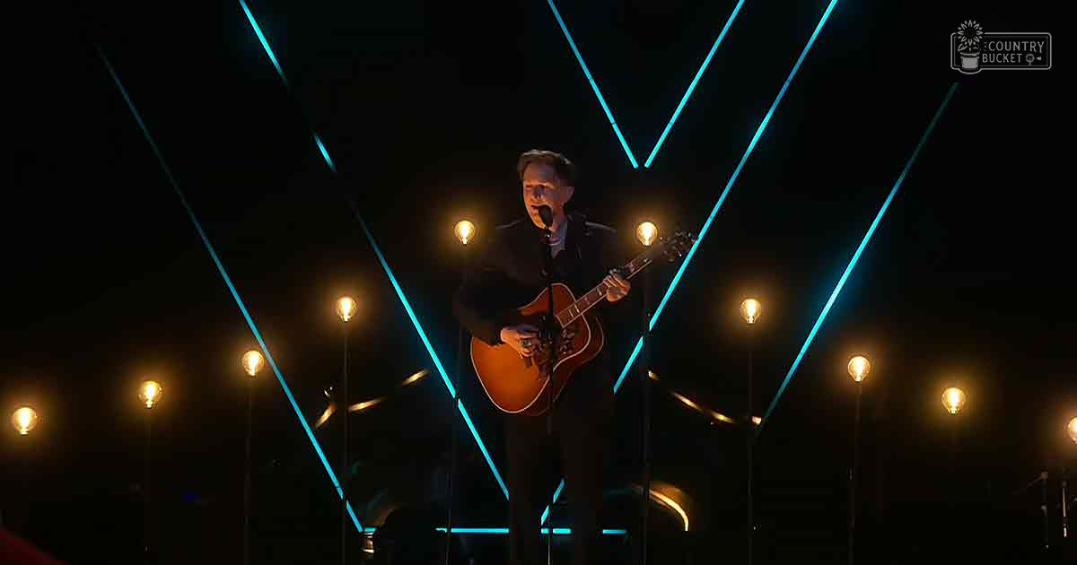 A male adult singer performing on a stage with warm yellow and cool blue lighting, wearing a dark suit.