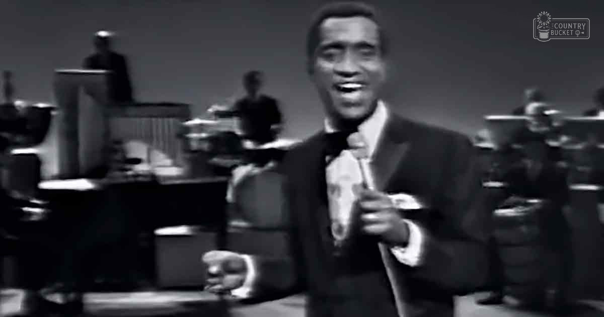 A male adult singer performing on stage with an orchestra behind him, dressed formally in a mid-20th-century dark suit and white shirt.