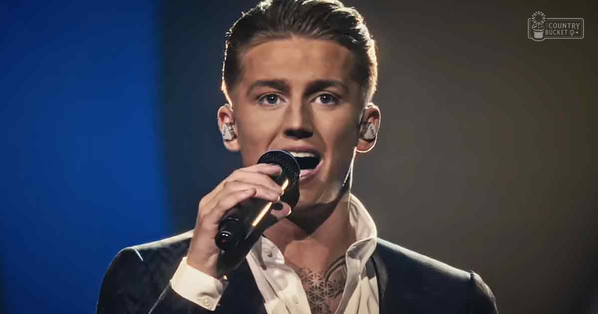 A young adult male performer on stage wearing a formal black suit with an open-collared white shirt, performing under professional stage lighting.