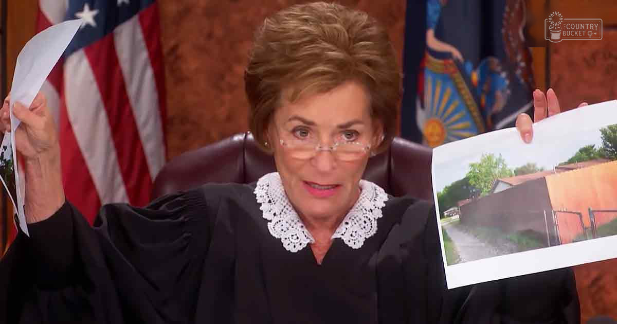 A middle-aged to older female judge in a formal black judicial robe with white lace trim, seated behind a dark leather chair in an American courtroom setting.
