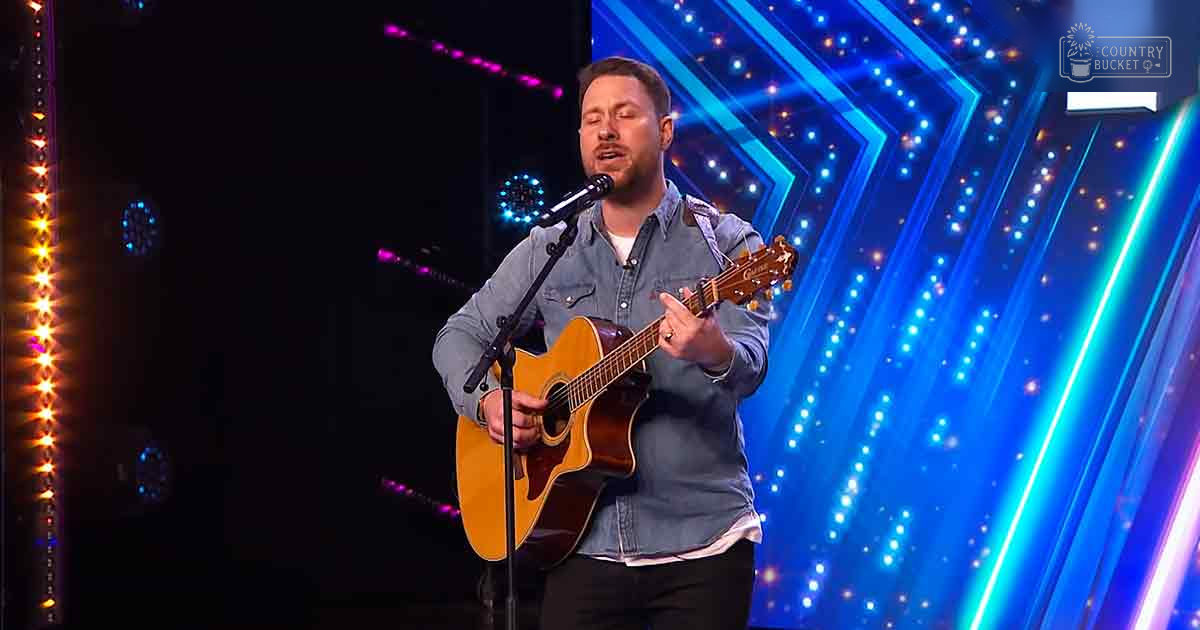 A male adult wearing a denim button-down shirt performing on stage under colorful stage lighting, with a guitar nearby.