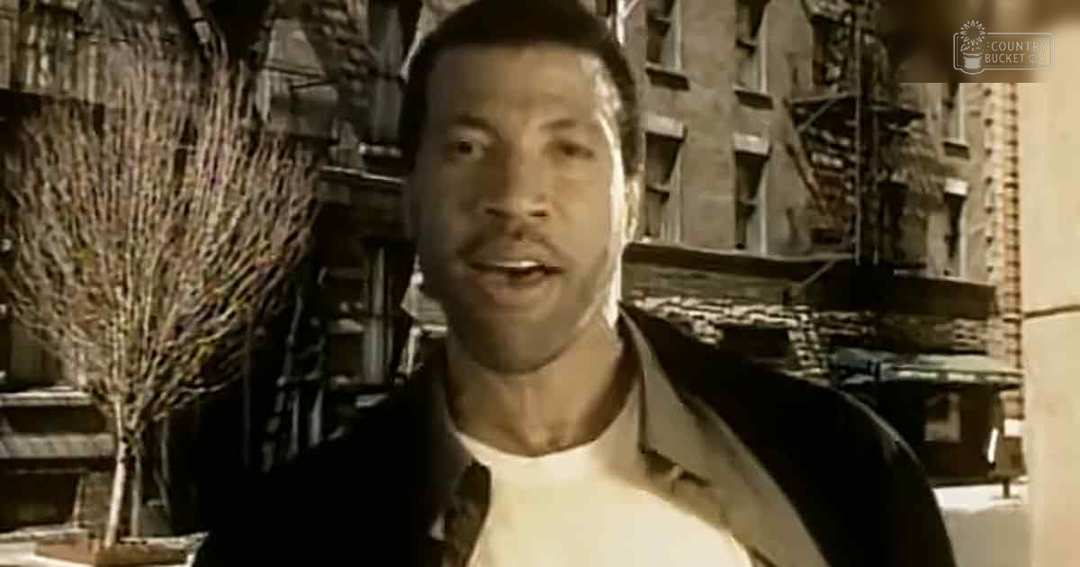 A young adult male stands in an urban alleyway with brick buildings and bare trees, wearing a relaxed black jacket over a white shirt.