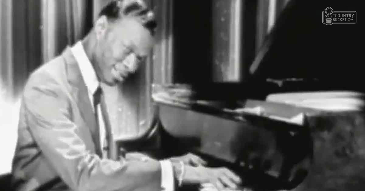 A middle-aged adult male in a light-colored suit jacket playing the grand piano in an indoor setting with curtains suggesting daytime.