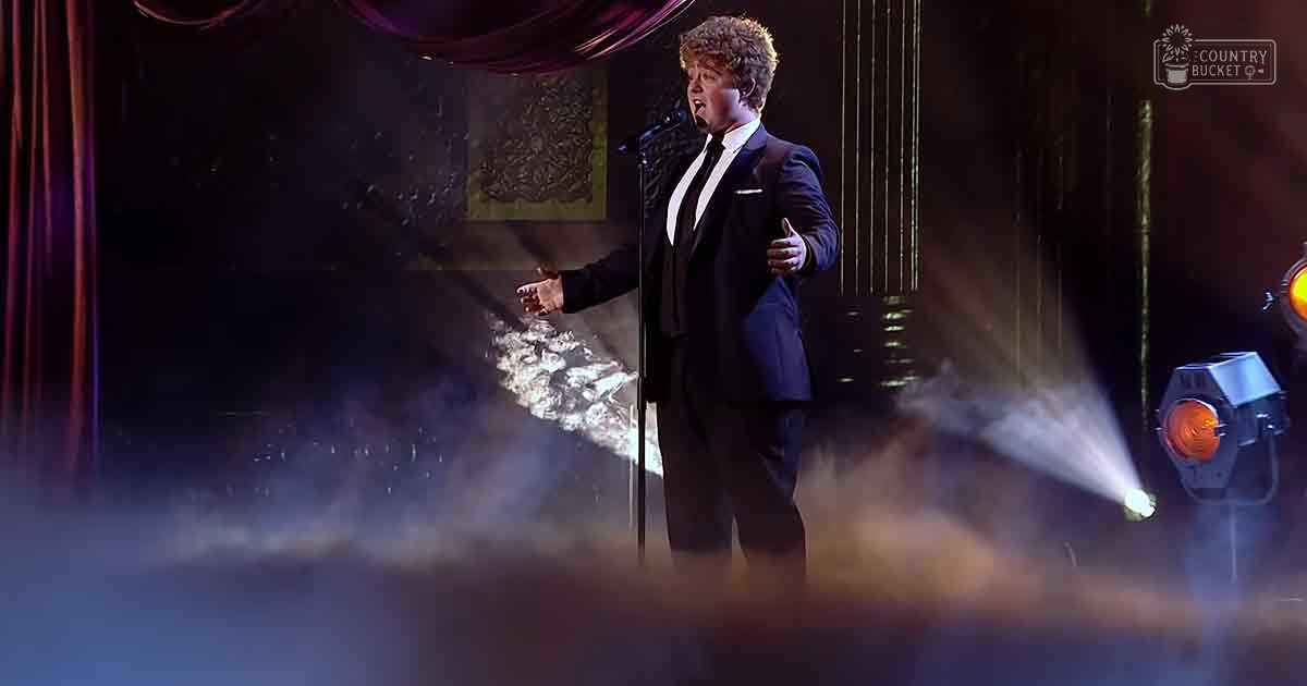 A young adult male performer in a tailored dark blue suit with a white shirt and black tie stands on stage, illuminated by an orange light beam against a backdrop of dark curtains and decorative panels. The setting suggests a formal event or performance space.