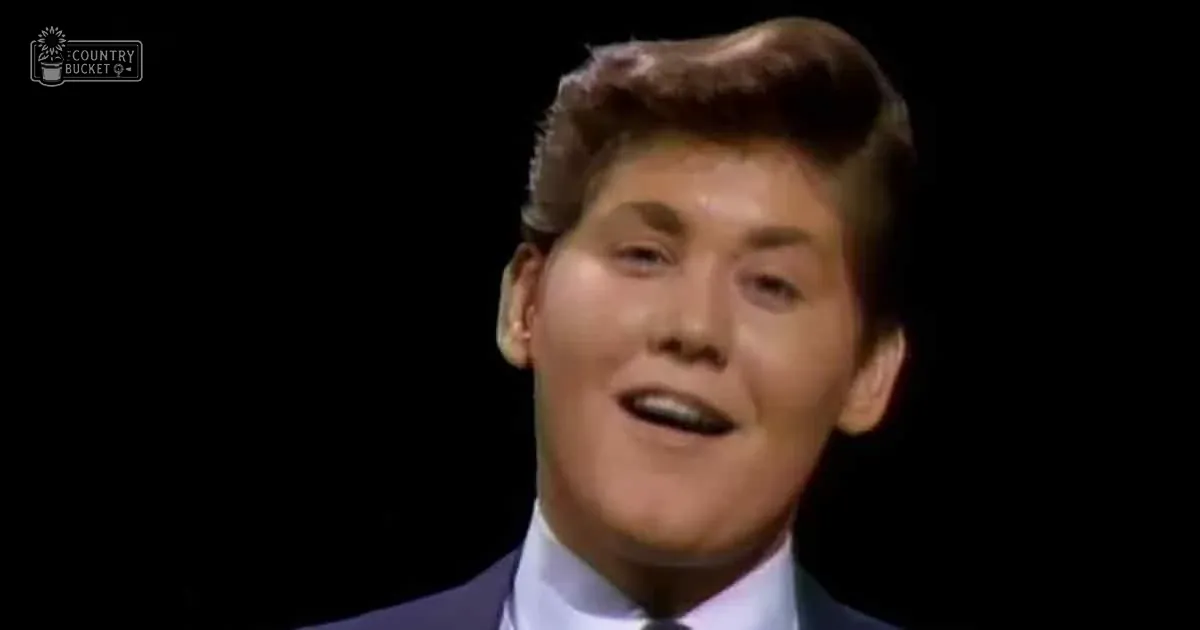 A young adult male with short, styled hair wears a light blue dress shirt under a dark suit jacket against a dark backdrop. Likely indoors during an event or performance.