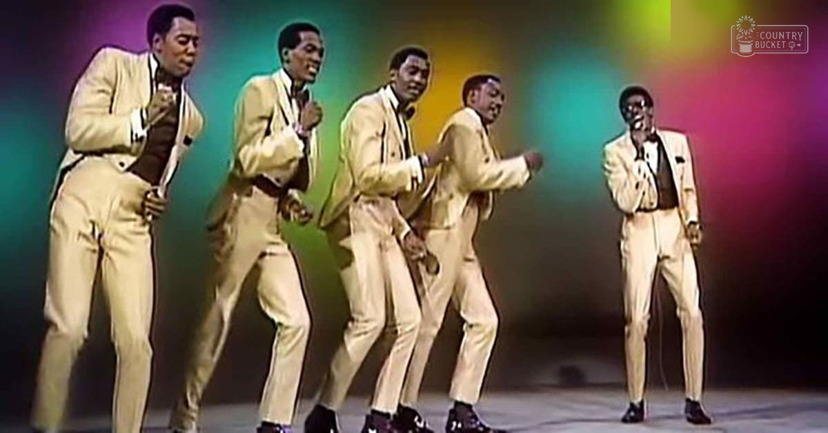 Adult males performing on an indoor stage with colorful lighting, wearing light-colored suits with dark vests and ties.