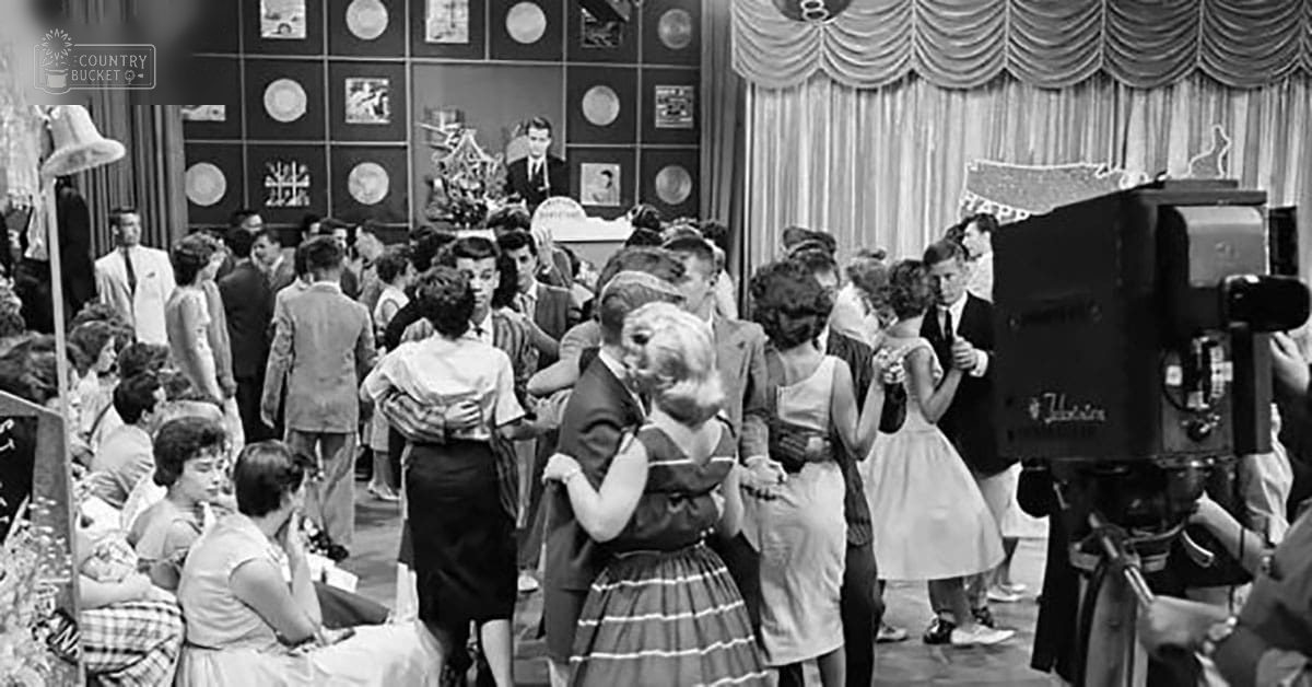A dance scene with teens/young adults in a studio setting, with mid-20th century-style dresses and suits under artificial lighting.
