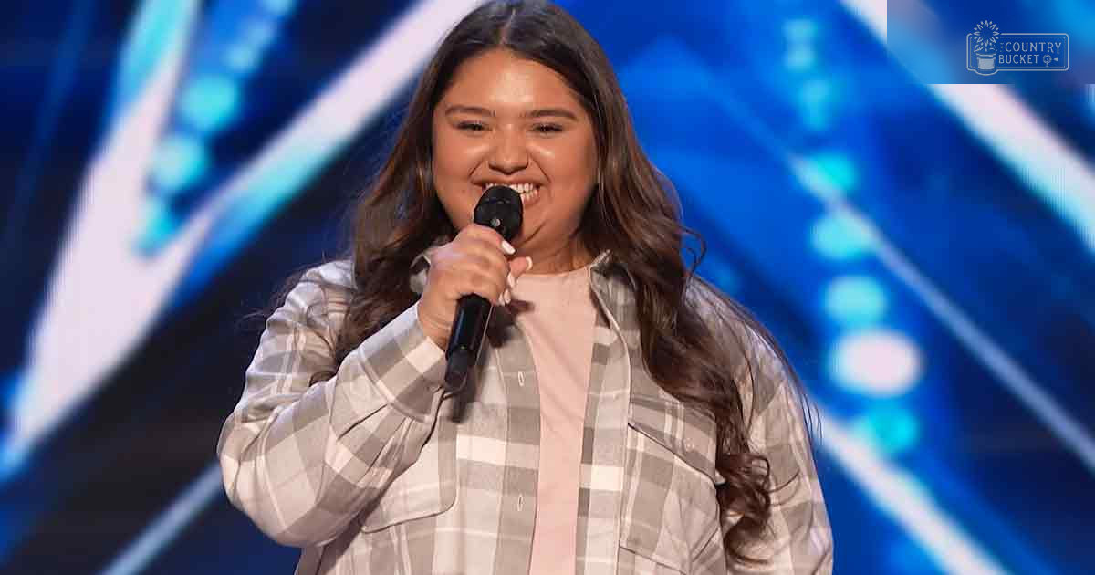 Young adult female wearing a plaid shirt over a pink collared shirt on stage with blue lighting.