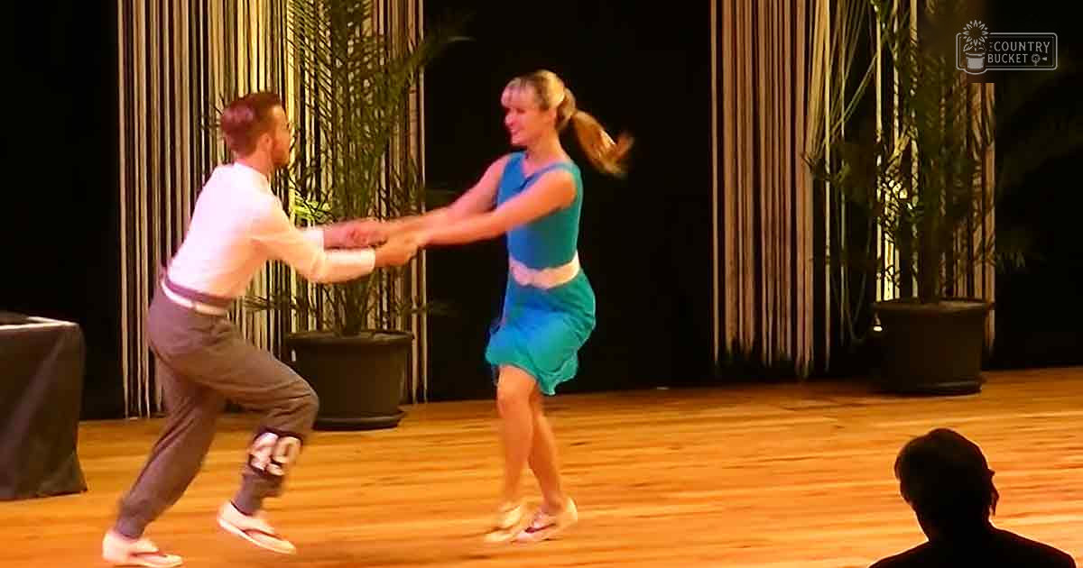 A dance performance in an indoor setting with two young adult dancers, one male wearing a white long-sleeved shirt and dark brown pants, and one female in a teal sleeveless dress, surrounded by wooden flooring, vertical blinds, potted plants, and warm stage lighting.