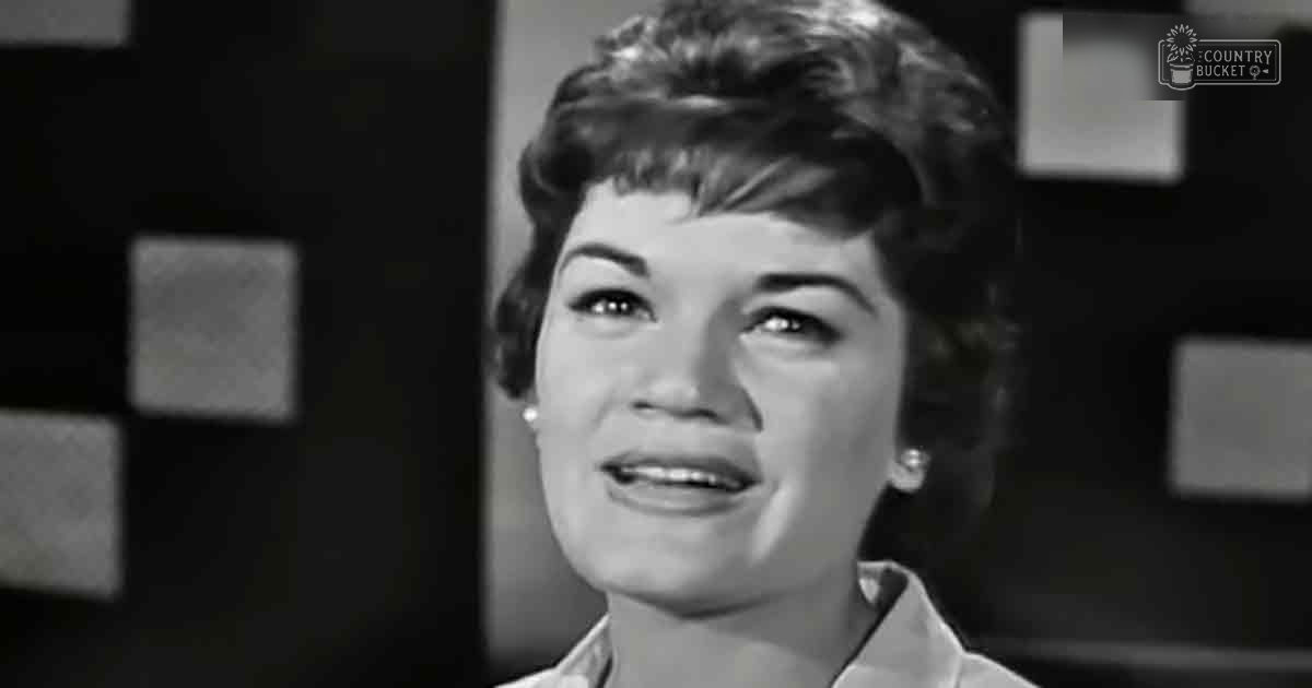 A middle-aged adult female wearing a mid-20th century-style collared shirt indoors with geometric patterns in the background.