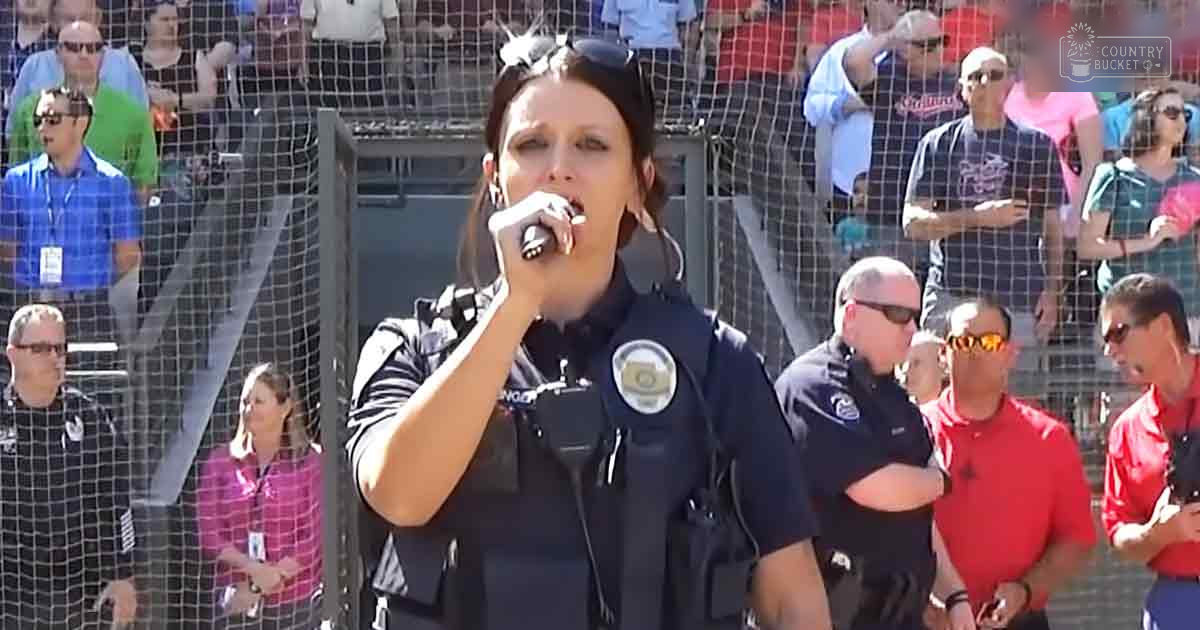 A woman singing at an outdoor event in a stadium-like setting with a crowd of spectators wearing summer attire, under bright daylight.