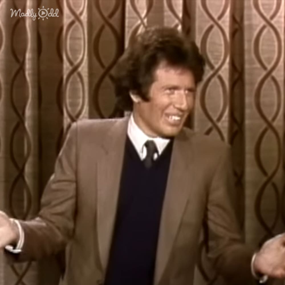 A middle-aged adult male in a relaxed fit brown blazer with a white shirt and dark tie, wearing a silver bracelet, indoors with patterned curtains.
