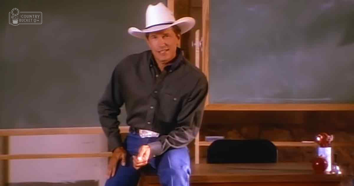 A middle-aged adult male wearing a long-sleeved button-up shirt with relaxed fit and blue jeans stands in an indoor setting resembling a classroom, surrounded by furniture such as a wooden desk with a black speaker and other small items.