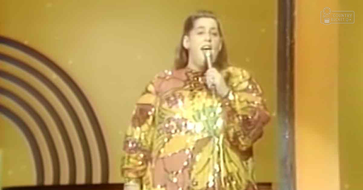 An adult female performer wearing a long-sleeved, sequined floral-patterned costume with gold accents on an indoor stage-like setting under warm 1970s-style lighting.