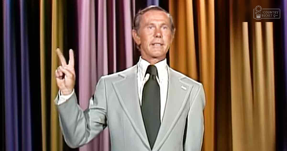 An older adult male in a formal setting on stage with purple and gold curtains.