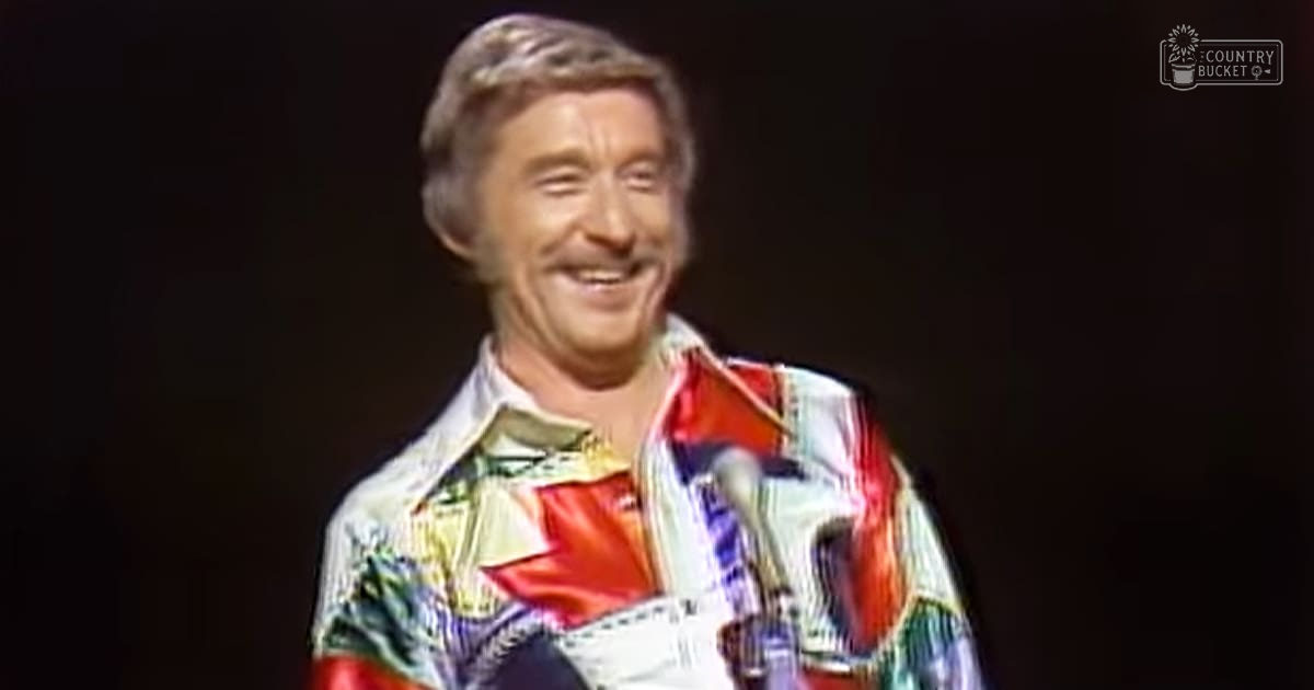 A middle-aged adult male wearing a relaxed, casual short-sleeved shirt with an abstract pattern resembling currency stands against a dark stage background under controlled lighting.