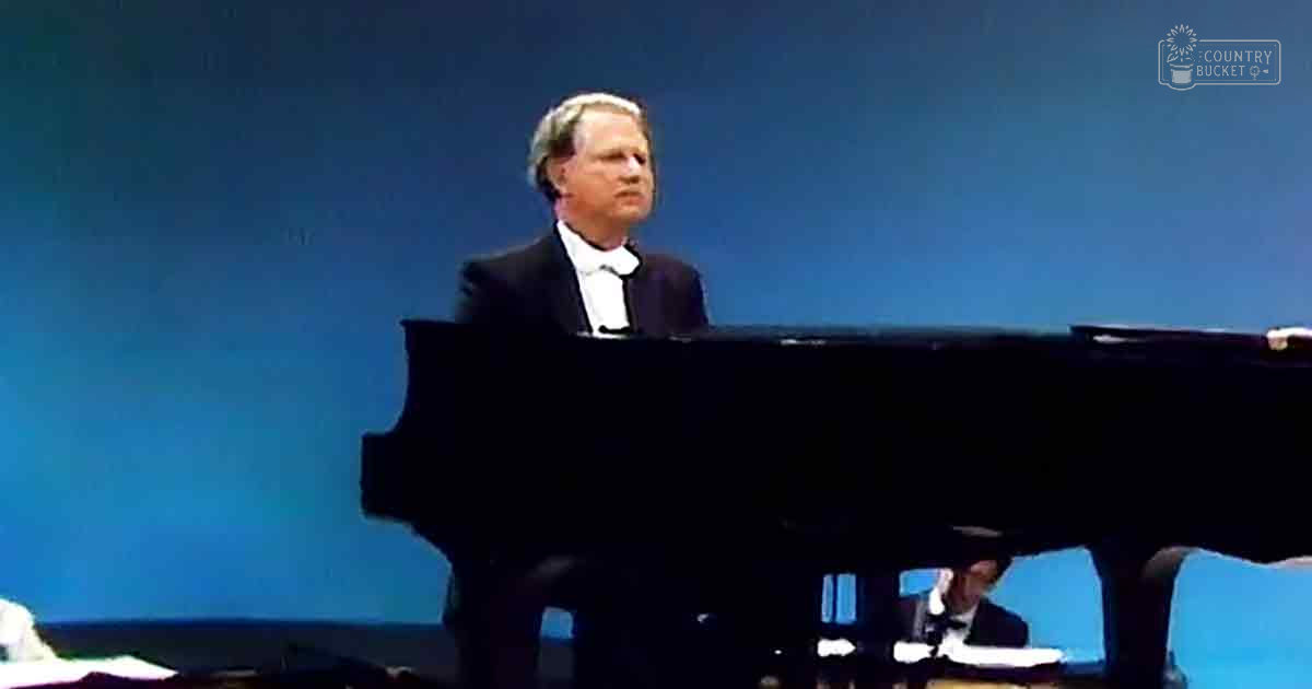 A middle-aged man in a dark suit and white shirt plays a grand piano in an indoor concert hall with stage lighting.