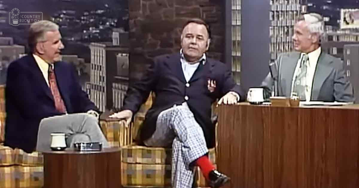 A middle-aged man seated in a television studio, surrounded by other adults dressed in formal attire; urban night scene backdrop suggests evening broadcast.