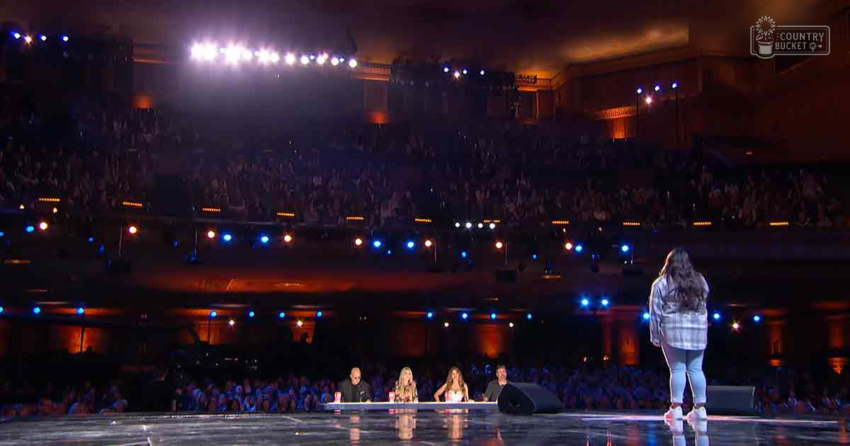 A young adult female contestant in a plaid shirt and light-colored pants stands on stage at an evening talent show, with judges seated behind her. An audience is visible in the background under artificial lighting.