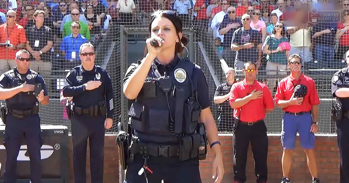 An adult female singer in a police uniform performs into a microphone at an outdoor stadium event, accompanied by several male officers in similar uniforms and another man in casual attire.