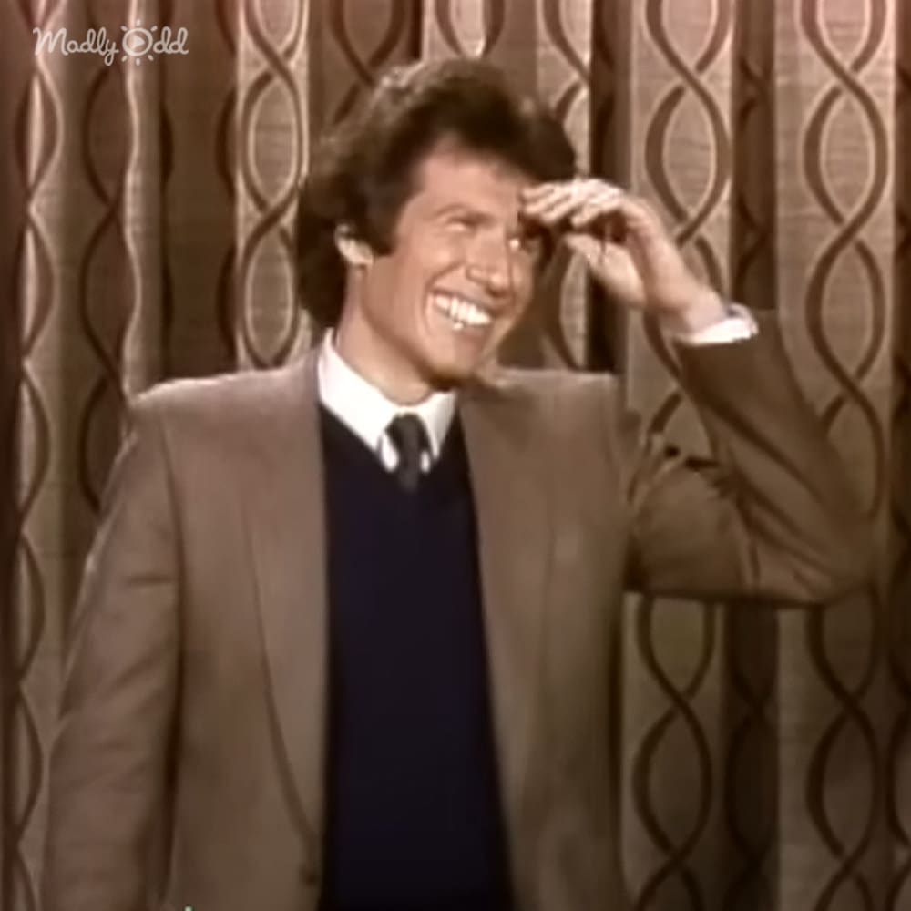 A middle-aged man wearing a light brown suit jacket and dark blue sweater vest in an indoor setting with patterned curtains, possibly for a talk show or performance.