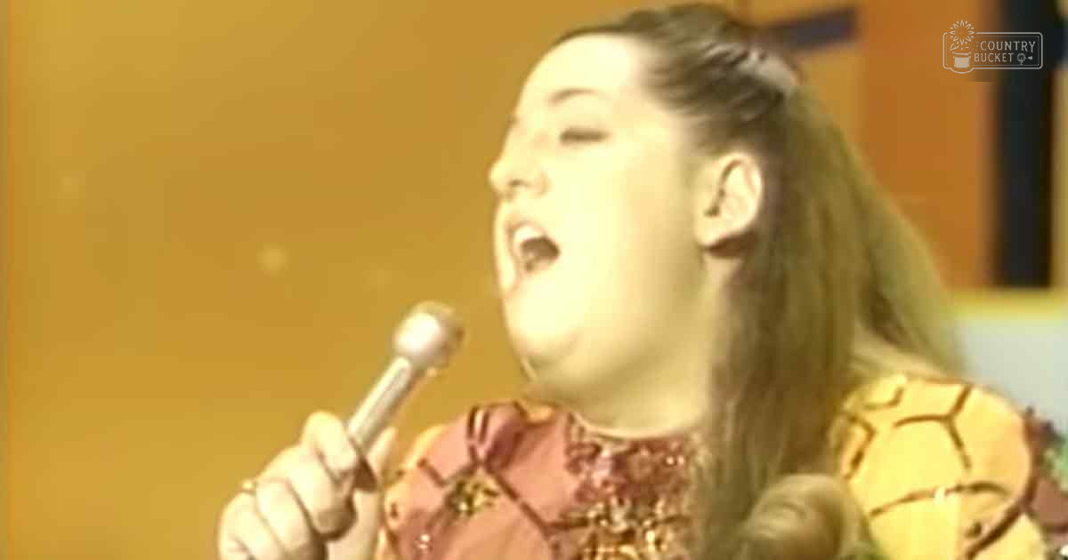 A young adult female singer in a traditional-style dress sings into a microphone on stage under warm lighting.