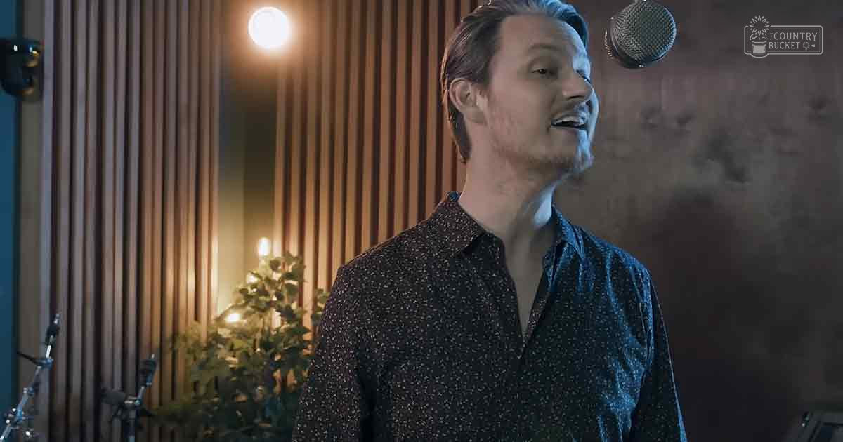 A young adult male in a recording studio setting, wearing a dark blue collared shirt with white polka dots and surrounded by wooden paneling and warm lighting.