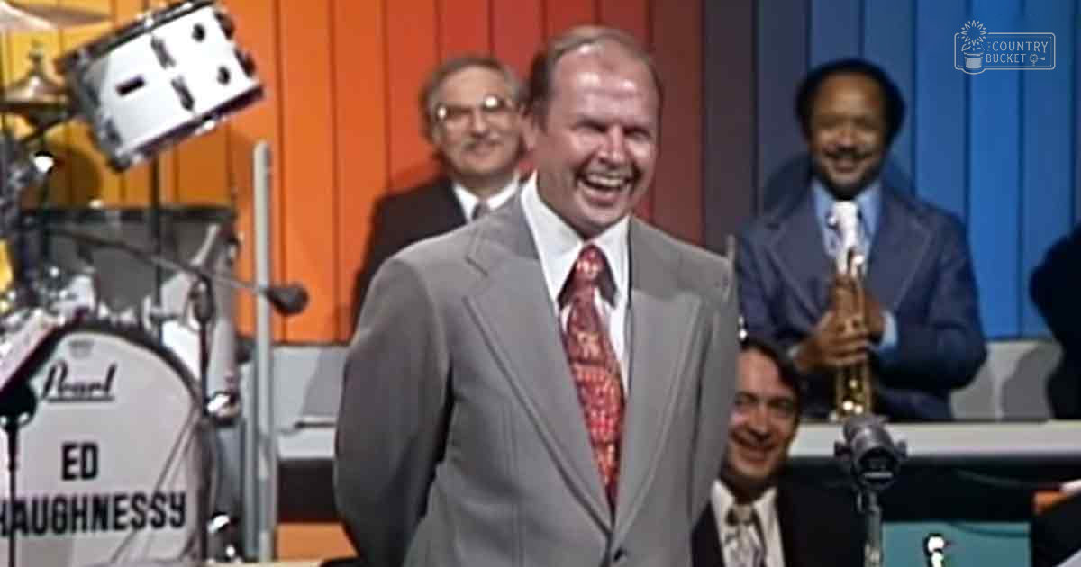 A middle-aged man in a gray suit with a red tie stands before a drum set, surrounded by other adults dressed formally in an indoor studio setting.