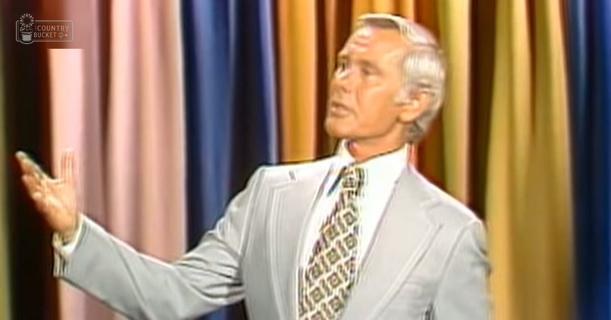 An older adult male wearing a light gray suit jacket with a subtle geometric-patterned tie against colorful striped curtains in what appears to be a professional studio setting.