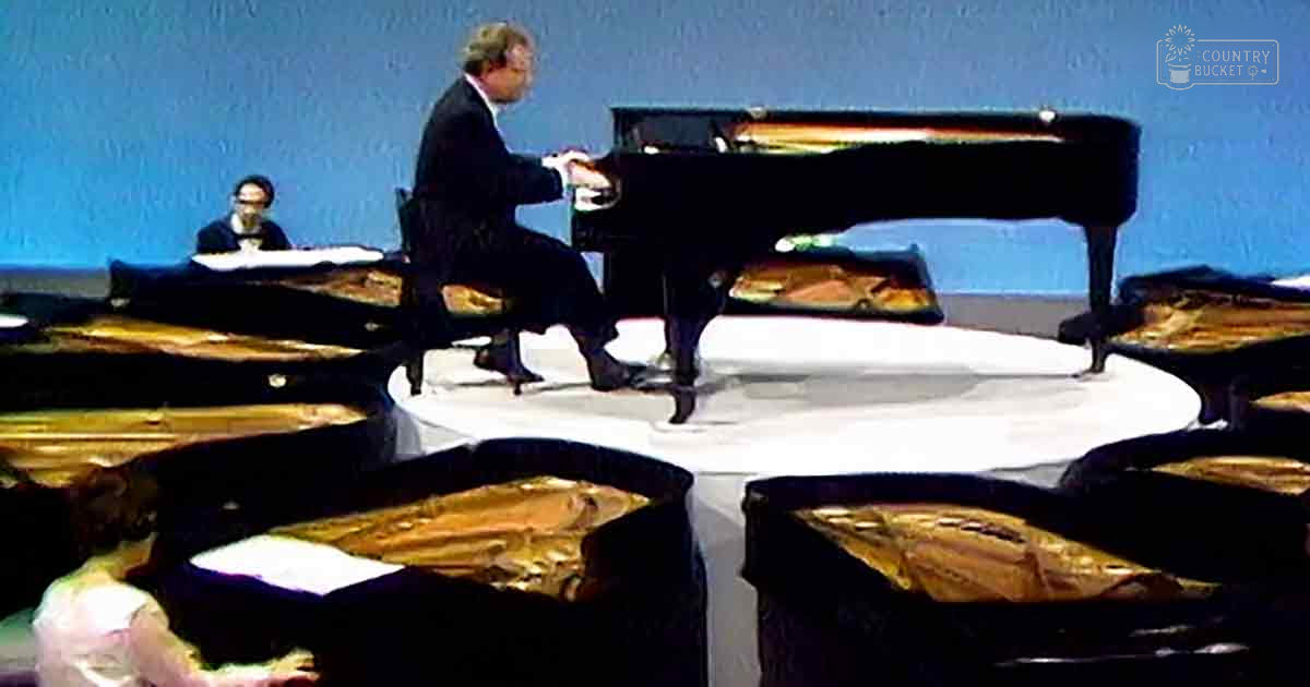 A middle-aged man with short hair and glasses plays a grand piano in an auditorium under artificial lighting.