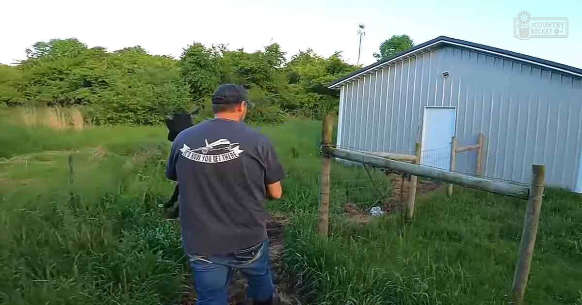 A young adult male wearing a dark gray t-shirt with graphic design on the back and front, standing in a rural outdoor setting with greenery around him.