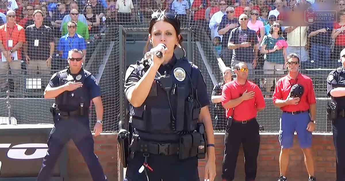 Adult female police officer singing at a public event with an audience behind a netted barrier, outdoors during daytime.