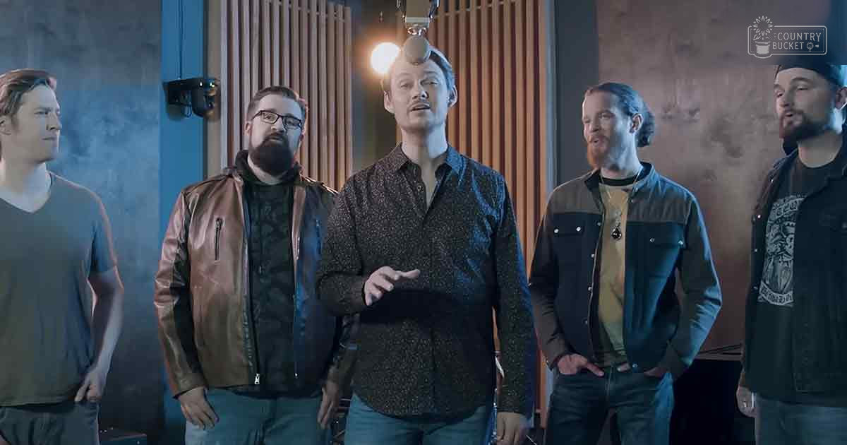 A group of adult males wearing casual attire stands indoors against wooden paneling with a textured wall, under artificial ceiling lights.