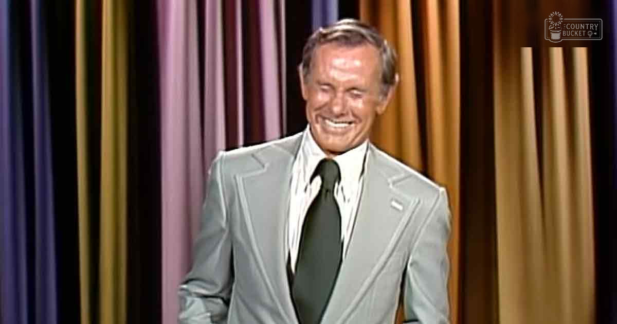 A middle-aged adult male wearing a formal light gray suit jacket, white shirt, and dark green tie stands in an indoor setting with curtains in purple, gold, and brown as the background.