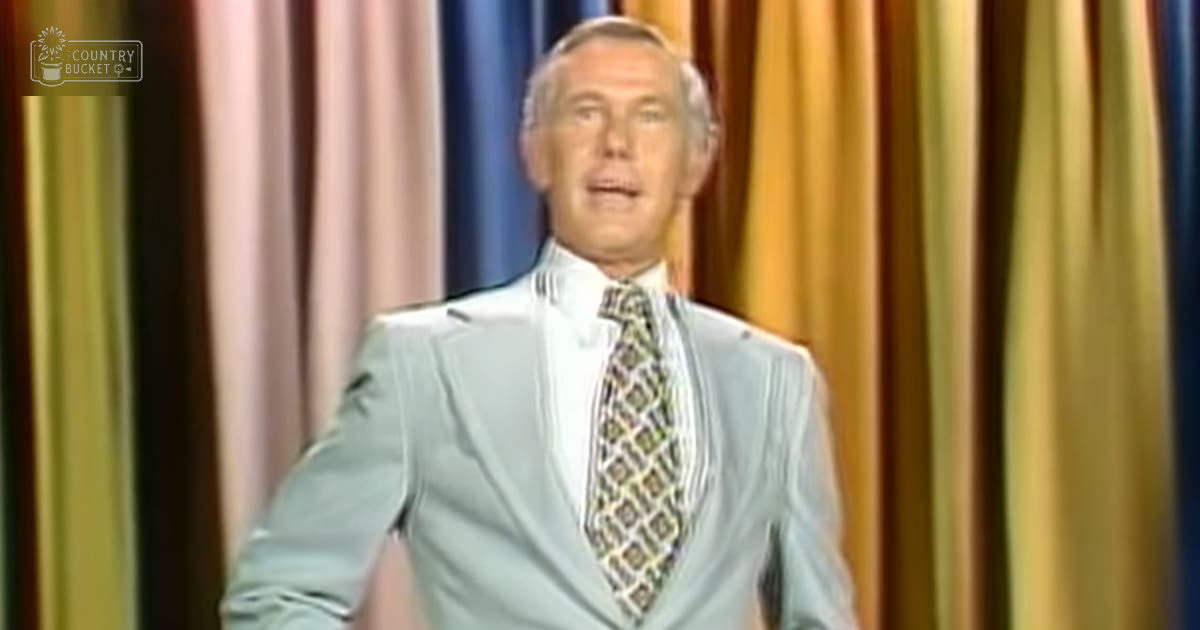 Older adult male in a studio setting with light blue suit jacket and white shirt with patterned tie with geometric design likely silk or polyester blend mid-20th century style, vertical striped curtain backdrop