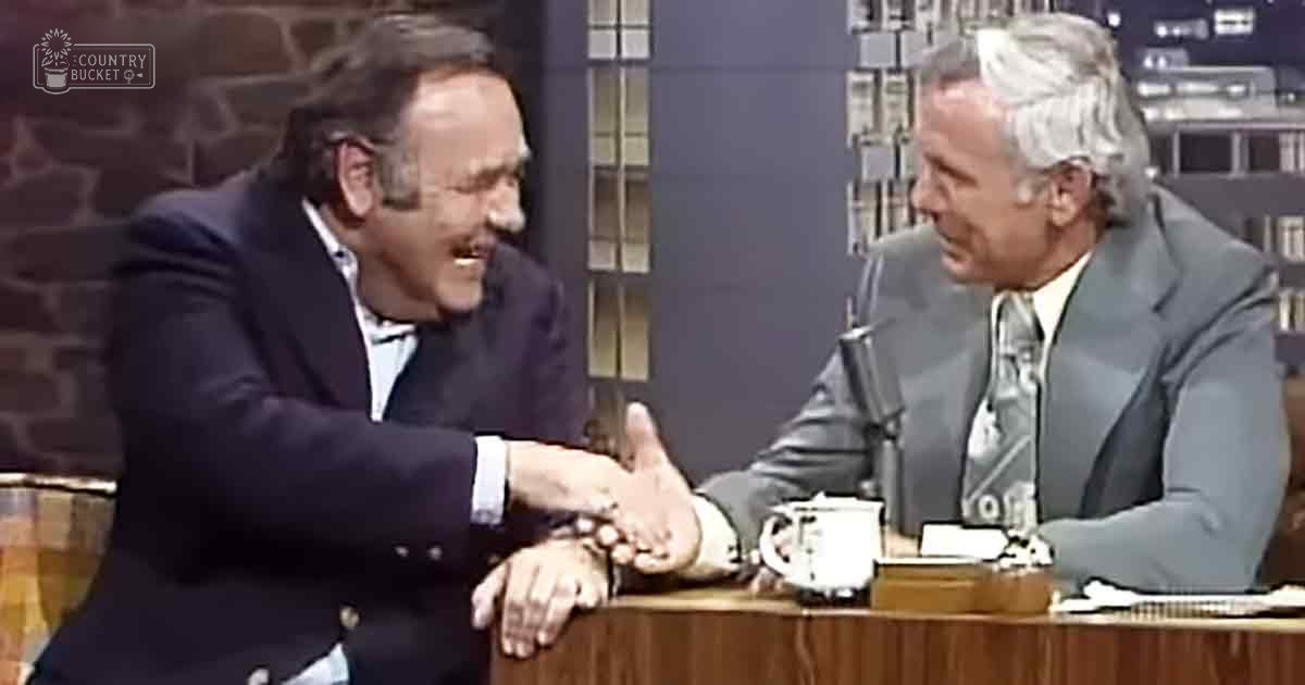 Two older adult men in a talk show setting, wearing classic mid-20th-century suits with patterned ties, seated at a wooden desk with a microphone and papers.