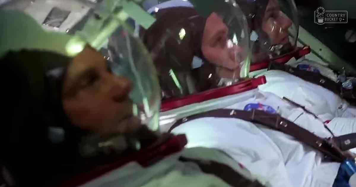 Astronauts in white spacesuits with red and black straps during what appears to be an EVA inside a spacecraft.