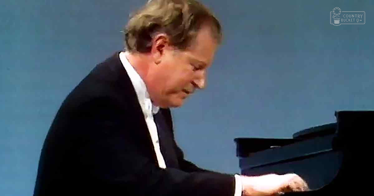 Middle-aged man playing a grand piano in an indoor performance venue with a plain blue backdrop, wearing a dark suit and white shirt.