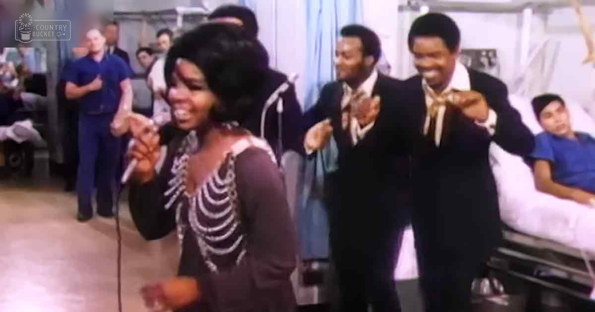 A young adult female sings into a microphone in what appears to be a hospital setting with several middle-aged men in formal attire and medical staff in the background.