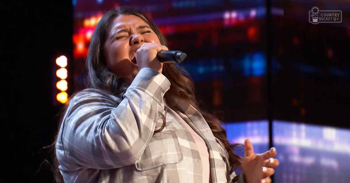A young adult female sings into a microphone during what appears to be a talent show, wearing a light-colored plaid shirt with long sleeves and button-up front.