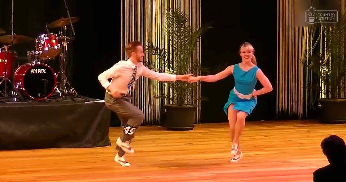 A dance performance scene with young adult male and female dancers in a formal indoor setting with stage lighting, curtains, and audience presence.
