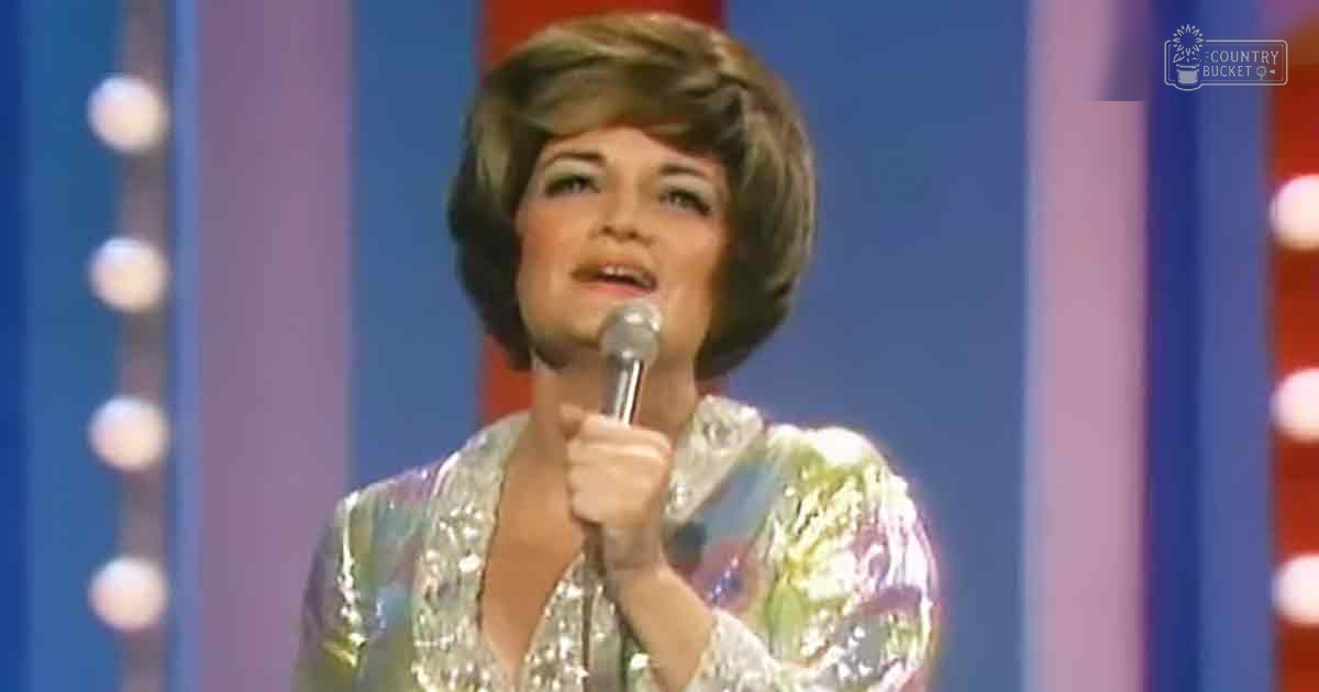 An adult female performer in a shiny, sequined 1960s or 1970s-style top holding a microphone against a stage backdrop with vertical stripes of red, white, and blue.