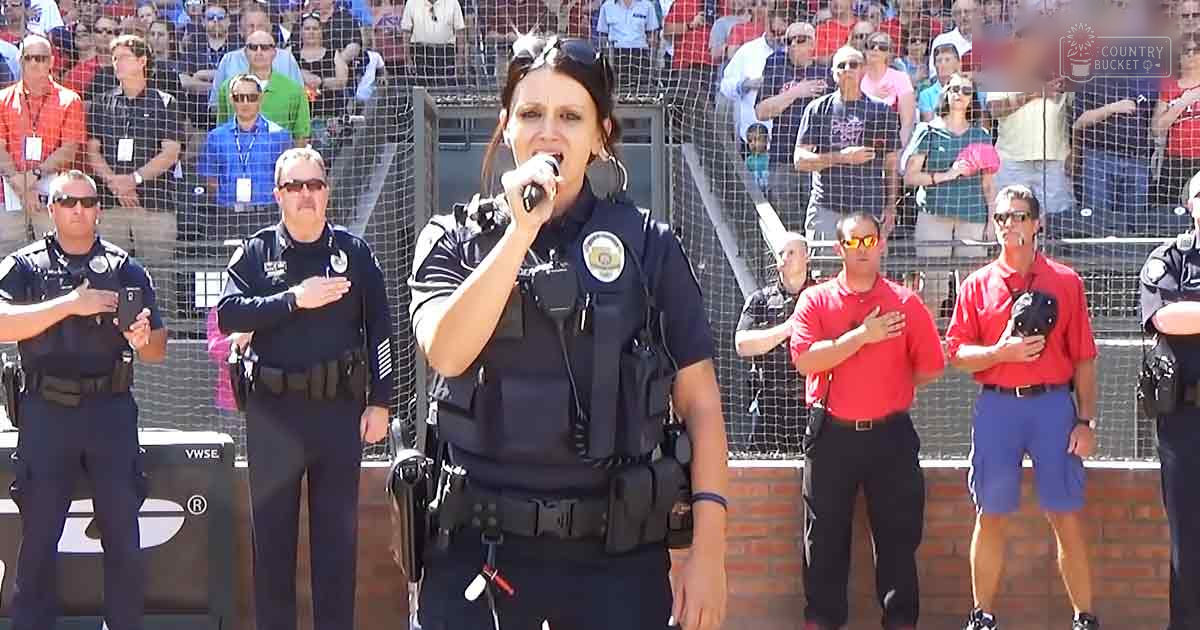 A woman in a black tactical vest sings into a microphone at an outdoor event with spectators.
