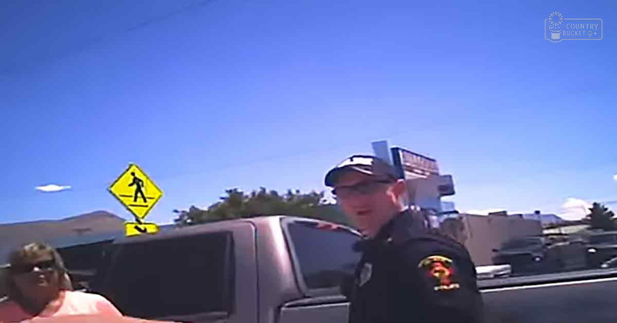 An adult male police officer and another individual stand outdoors under clear blue skies, near a dark-colored pickup truck in an urban environment.